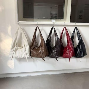 Puede incluir: Cinco bolsos de hombro de cuero en varios colores: blanco, marr&oacute;n, marr&oacute;n oscuro, rojo y negro. Cada bolso tiene forma de media luna y est&aacute; suspendido de un gancho de metal. Los bolsos tienen una textura suave y brillante.