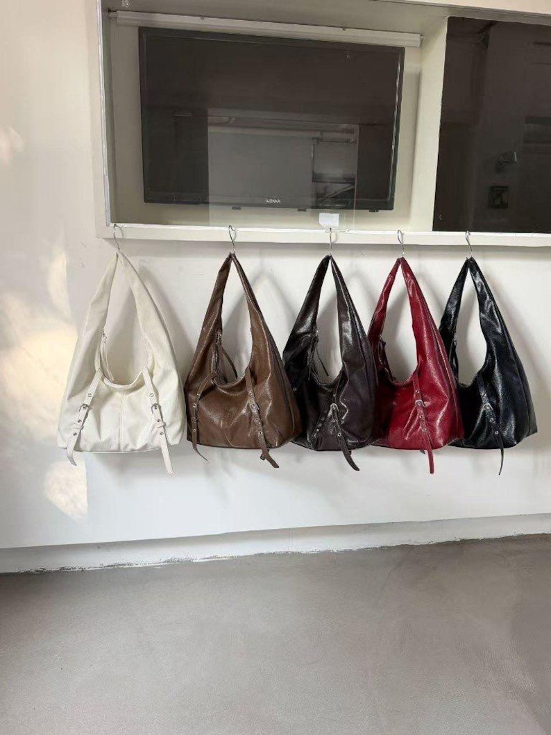 Puede incluir: Cinco bolsos de hombro de cuero en varios colores: blanco, marr&oacute;n, marr&oacute;n oscuro, rojo y negro. Cada bolso tiene forma de media luna y est&aacute; suspendido de un gancho de metal. Los bolsos tienen una textura suave y brillante.