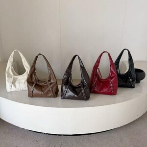 Puede incluir: Cinco bolsos de hombro de cuero en fila, cada uno de un color diferente. Los colores son blanco, marr&oacute;n, marr&oacute;n oscuro, rojo y negro. Cada bolso tiene forma curva y una sola correa para el hombro. Los bolsos est&aacute;n sobre una superficie blanca y curva.