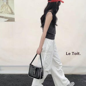 May include: A person wearing a red baseball cap, black vest top, and white trousers, carrying a black shoulder bag with white trim. The bag has a silver buckle. The text "Le Toit." is visible.