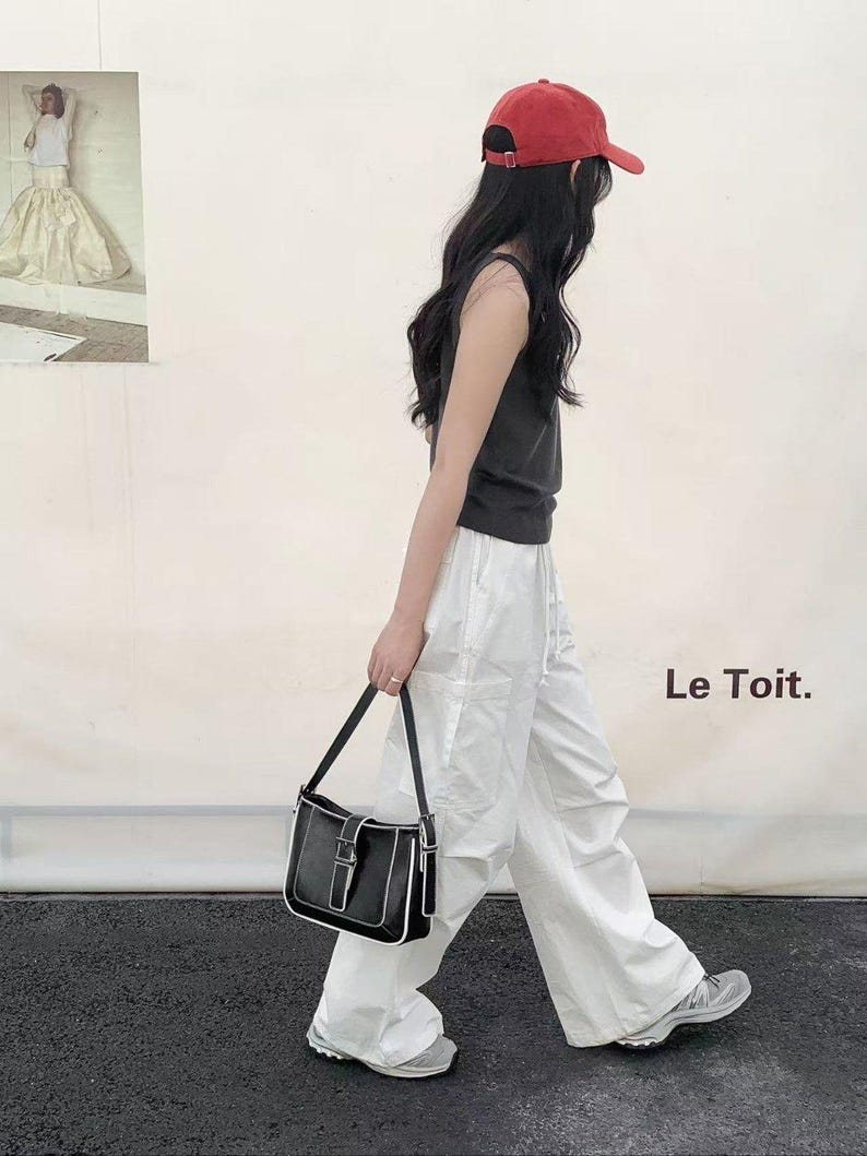 May include: A person wearing a red baseball cap, black vest top, and white trousers, carrying a black shoulder bag with white trim. The bag has a silver buckle. The text "Le Toit." is visible.