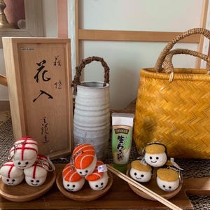 May include: A display of sushi-themed keyrings and accessories. The keyrings depict smiling sushi characters with red, orange, and yellow details. A wooden box with Japanese script, a wasabi tube, and a woven basket are also visible.