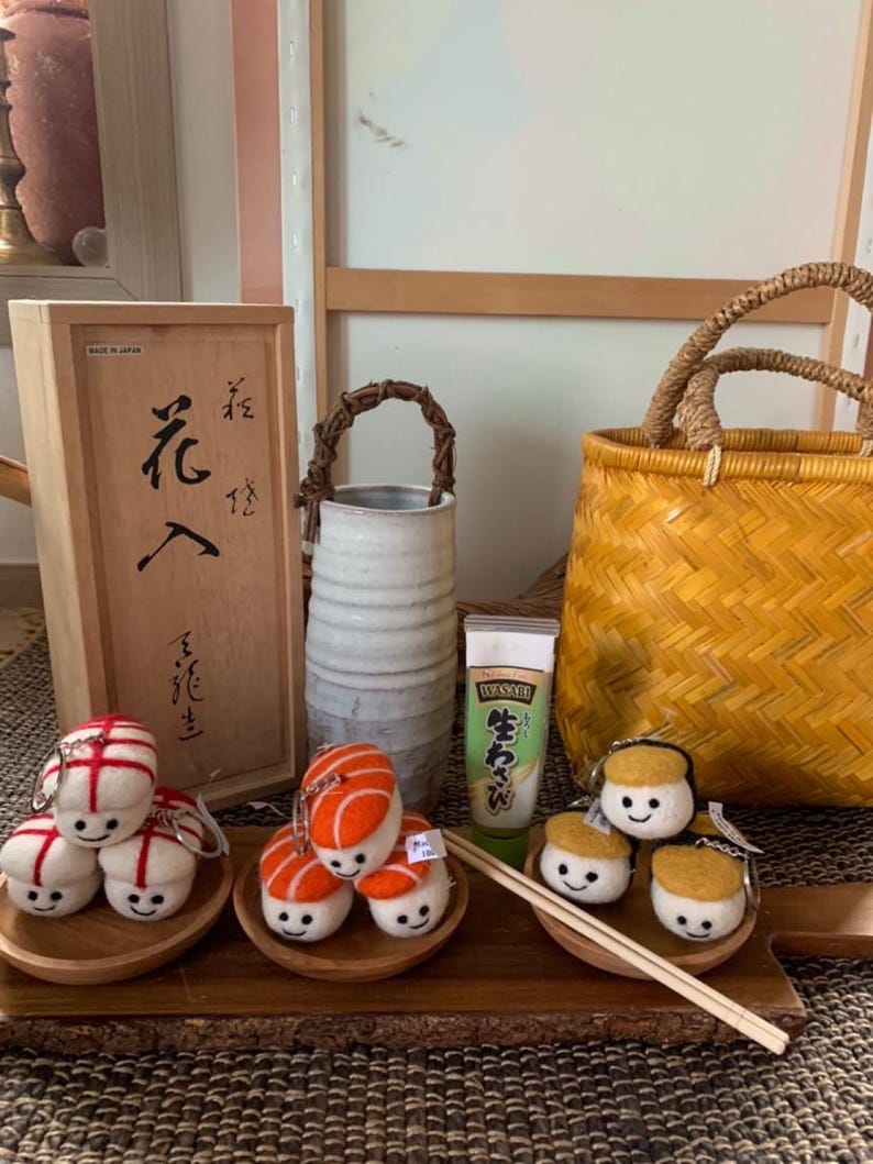 May include: A display of sushi-themed keyrings and accessories. The keyrings depict smiling sushi characters with red, orange, and yellow details. A wooden box with Japanese script, a wasabi tube, and a woven basket are also visible.