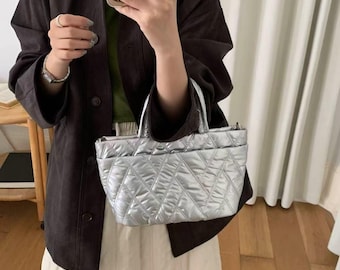 May include: A silver quilted tote bag with a diamond pattern. The bag has two small pockets on the front and two handles. The bag is held by a person wearing a dark brown jacket and a white skirt.