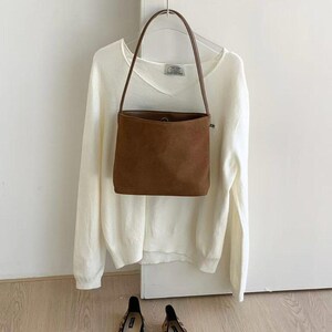May include: A brown suede handbag with a long handle hangs in front of a white sweater on a hanger. Below, a pair of leopard print flats with black bows sit on a wooden floor. The scene is set against a white wall.
