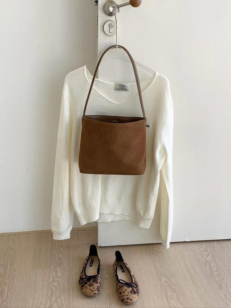 May include: A brown suede handbag with a long handle hangs in front of a white sweater on a hanger. Below, a pair of leopard print flats with black bows sit on a wooden floor. The scene is set against a white wall.