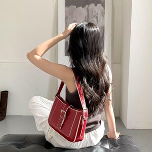 May include: A red shoulder bag with a rectangular shape and a silver buckle. The bag has a white trim and a matching strap. The person is wearing a brown top and white trousers.