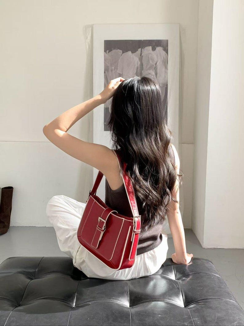May include: A red shoulder bag with a rectangular shape and a silver buckle. The bag has a white trim and a matching strap. The person is wearing a brown top and white trousers.