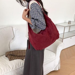 May include: A burgundy suede shoulder bag with a long strap. The bag is worn over the shoulder. The person is wearing a plaid shirt, gray pants, and a gray beret. A white couch is in the background.