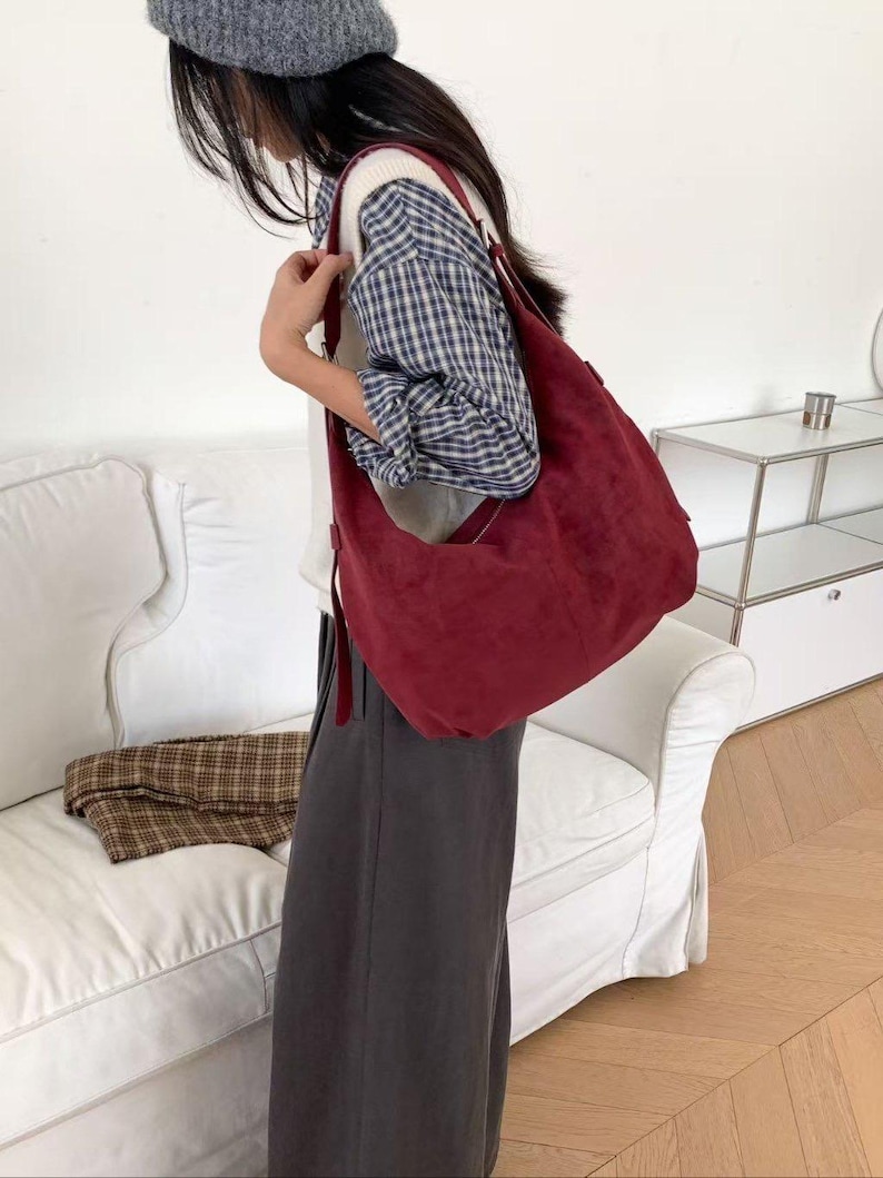 May include: A burgundy suede shoulder bag with a long strap. The bag is worn over the shoulder. The person is wearing a plaid shirt, gray pants, and a gray beret. A white couch is in the background.