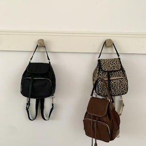 Puede incluir: Tres mochilas colgadas en una pared blanca. Una es negra, otra con estampado de leopardo y la tercera marrón. Todas tienen correas ajustables y están suspendidas de pequeños ganchos redondos.