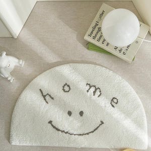 Puede incluir: Una alfombra de baño blanca semicircular con la palabra "home" y una cara sonriente bordada en gris. La alfombra está sobre una superficie de tonos neutros, con una pequeña lámpara y libros en el fondo.