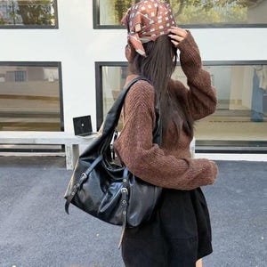 Puede incluir: Un bolso de hombro de cuero negro con herrajes plateados. El bolso tiene un dise&ntilde;o holgado con m&uacute;ltiples correas y hebillas. La persona lleva un su&eacute;ter marr&oacute;n, una falda negra y un pa&ntilde;uelo estampado.