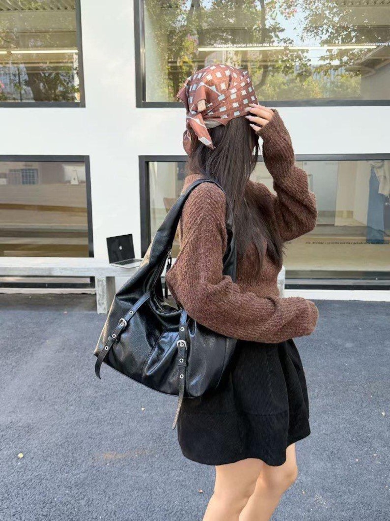 Puede incluir: Un bolso de hombro de cuero negro con herrajes plateados. El bolso tiene un dise&ntilde;o holgado con m&uacute;ltiples correas y hebillas. La persona lleva un su&eacute;ter marr&oacute;n, una falda negra y un pa&ntilde;uelo estampado.