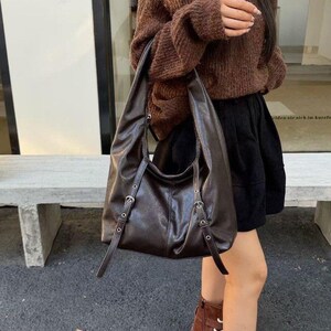 Puede incluir: Un bolso de hombro de cuero marr&oacute;n oscuro con correas ajustables y hebillas plateadas. El bolso tiene un dise&ntilde;o holgado y una sola correa para el hombro. El bolso lo lleva una persona que lleva un su&eacute;ter marr&oacute;n y una falda negra.