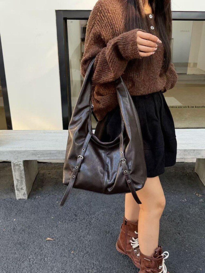 Puede incluir: Un bolso de hombro de cuero marr&oacute;n oscuro con correas ajustables y hebillas plateadas. El bolso tiene un dise&ntilde;o holgado y una sola correa para el hombro. El bolso lo lleva una persona que lleva un su&eacute;ter marr&oacute;n y una falda negra.