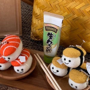 May include: A selection of sushi-themed keychains and a tube of wasabi are displayed. The keychains feature felt sushi pieces with smiling faces, including salmon nigiri and yellow-topped sushi. Chopsticks are placed on a small wooden plate.