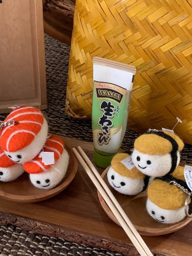 May include: A selection of sushi-themed keychains and a tube of wasabi are displayed. The keychains feature felt sushi pieces with smiling faces, including salmon nigiri and yellow-topped sushi. Chopsticks are placed on a small wooden plate.