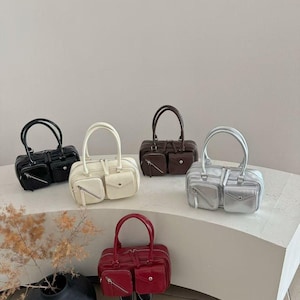 May include: A collection of six rectangular handbags in various colors: black, cream, brown, silver, and red. Each bag features two front pockets with zippers or snap closures and a top handle. The bags are displayed on a white surface.