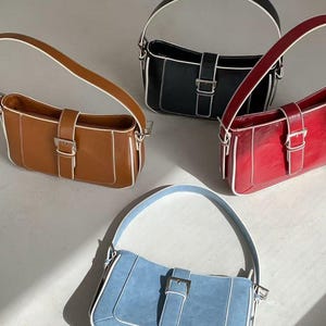 May include: Four rectangular shoulder bags in brown, black, red, and blue. Each bag has a silver buckle, white trim, and a matching handle. The bags are displayed on a light-coloured surface.