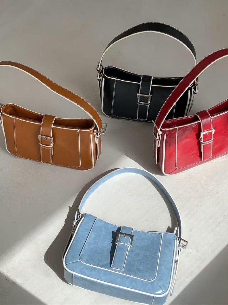 May include: Four rectangular shoulder bags in brown, black, red, and blue. Each bag has a silver buckle, white trim, and a matching handle. The bags are displayed on a light-coloured surface.