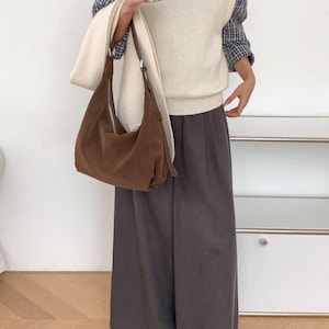 May include: A woman wearing a cream-colored sweater vest over a blue and white plaid shirt, paired with gray wide-leg pants and brown shoes. She carries a brown suede shoulder bag and a stainless steel travel mug. A cream-colored scarf is draped over the bag.