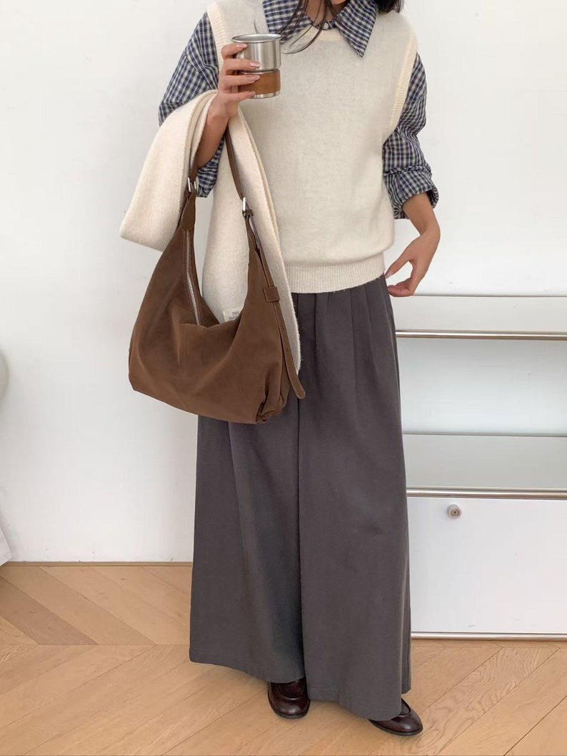 May include: A woman wearing a cream-colored sweater vest over a blue and white plaid shirt, paired with gray wide-leg pants and brown shoes. She carries a brown suede shoulder bag and a stainless steel travel mug. A cream-colored scarf is draped over the bag.