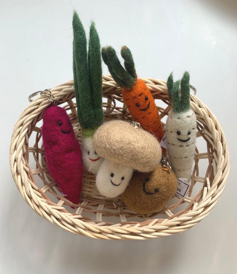 May include: A wicker basket filled with felt food keychains. The keychains are shaped like a carrot, mushroom, onion, potato, and a brown round object. Each keychain has a smiling face.