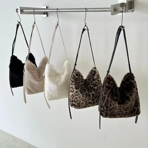 May include: Five faux fur shoulder bags in black, beige, white, and two leopard print patterns hang from a metal bar. Each bag has a long, thin black strap. The bags are displayed against a white wall.