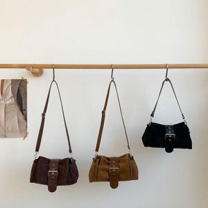 May include: Three suede shoulder bags in shades of brown and black hang from a wooden rod. Each bag features a rectangular shape, adjustable strap, and a large buckle closure. The bags are displayed against a plain white background.