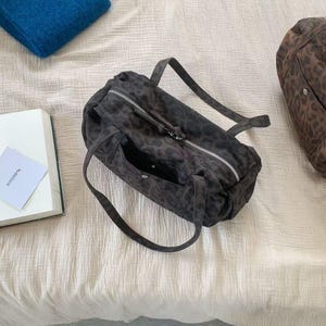 Puede incluir: Un bolso gris oscuro con estampado de leopardo, cremallera plateada y dos asas. Tambi&eacute;n se ven una manta azul doblada y un libro. El bolso mide aproximadamente 30 cm de largo.