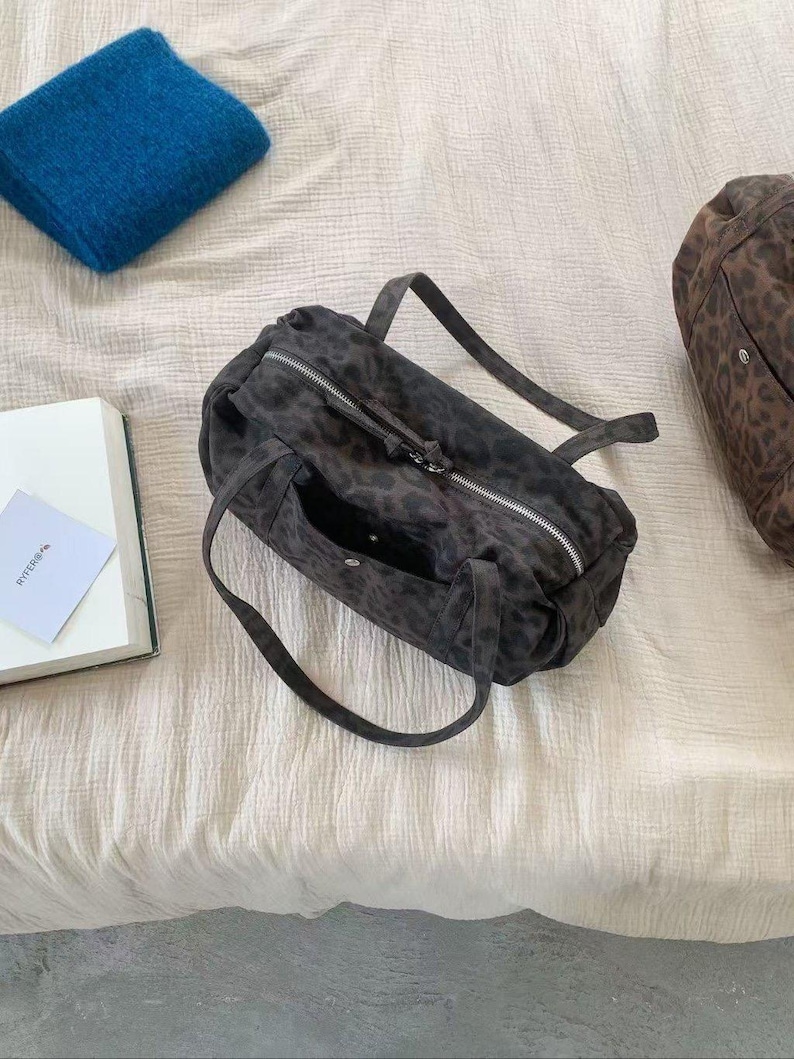 Puede incluir: Un bolso gris oscuro con estampado de leopardo, cremallera plateada y dos asas. Tambi&eacute;n se ven una manta azul doblada y un libro. El bolso mide aproximadamente 30 cm de largo.
