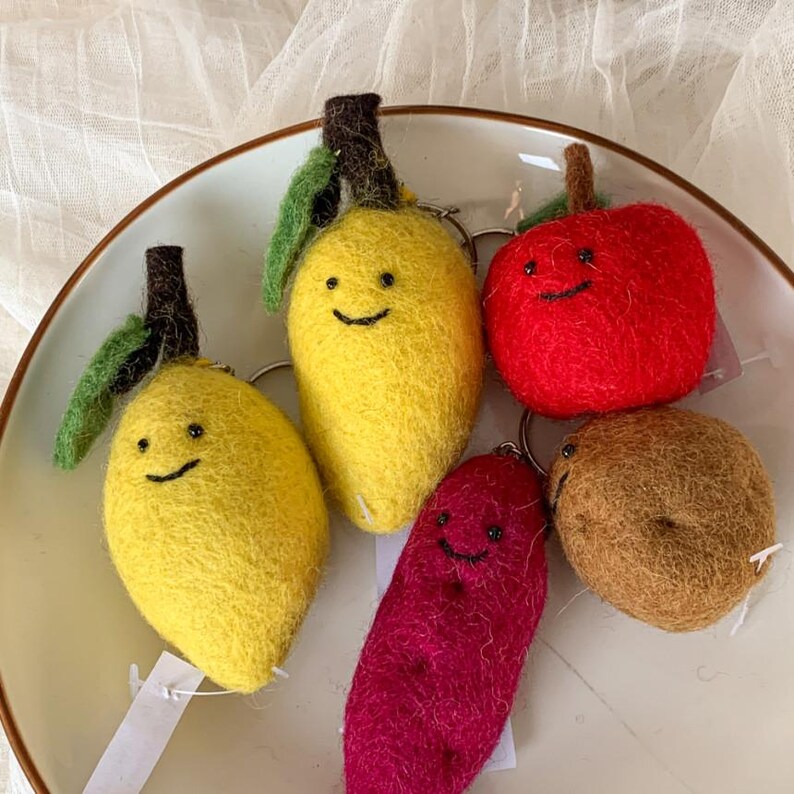 May include: Five felt fruit keychains with happy faces. The keychains are a yellow lemon, a yellow mango, a red apple, a brown potato, and a purple sweet potato.