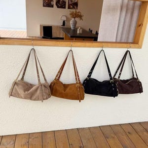 May include: Four suede shoulder bags in varying colors: tan, brown, black, and dark brown. Each bag has a rectangular shape with a long, thin strap and metal stud details. The bags are hanging on hooks against a white wall.