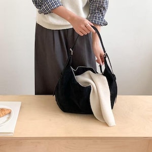 May include: A black suede shoulder bag with a curved design, held by a person. The bag is open, revealing a cream-colored folded sweater. The bag has a zipper closure and a long strap.