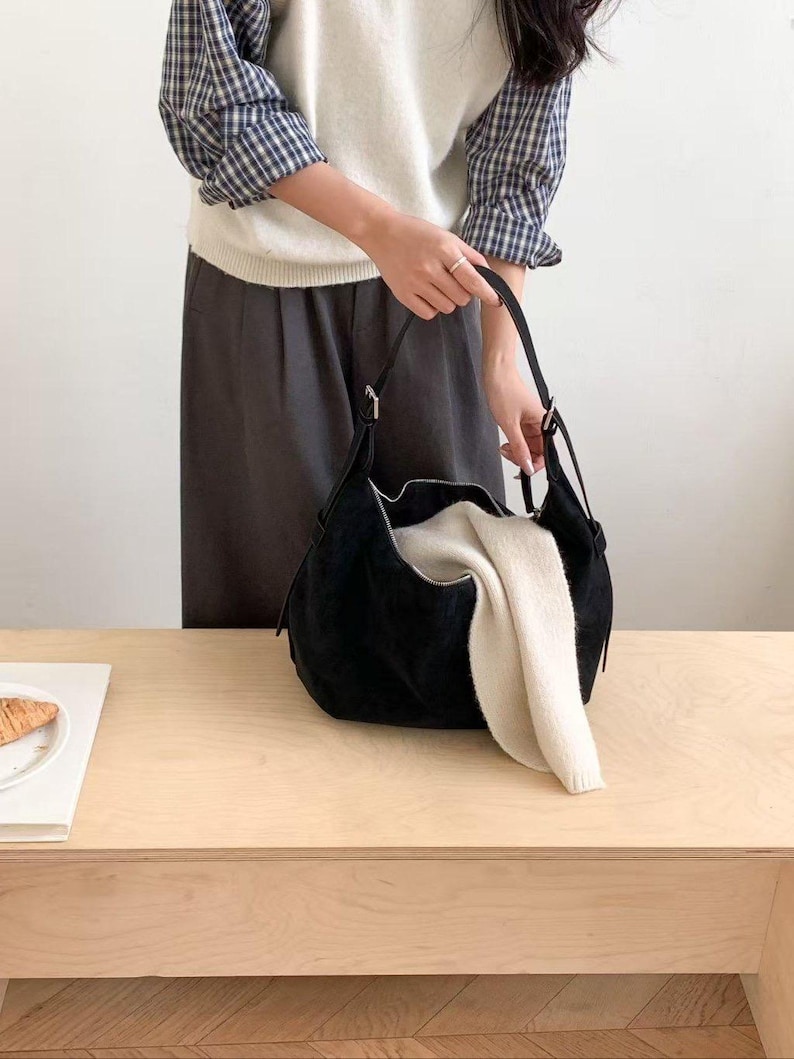 May include: A black suede shoulder bag with a curved design, held by a person. The bag is open, revealing a cream-colored folded sweater. The bag has a zipper closure and a long strap.