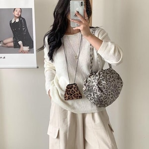 Puede incluir: Una persona con un su&eacute;ter blanco y pantalones beige, sosteniendo un bolso con estampado de leopardo y una peque&ntilde;a bolsa con estampado de leopardo. Un p&oacute;ster con una foto de una mujer con un abrigo negro est&aacute; en el fondo.