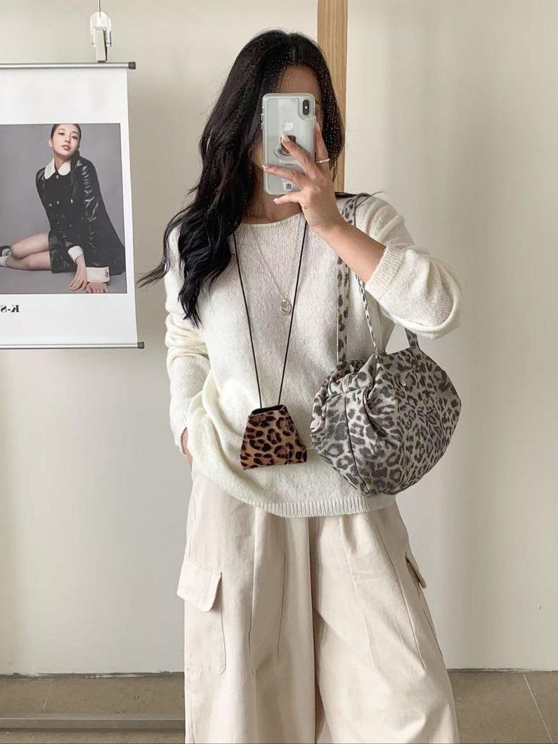 Puede incluir: Una persona con un su&eacute;ter blanco y pantalones beige, sosteniendo un bolso con estampado de leopardo y una peque&ntilde;a bolsa con estampado de leopardo. Un p&oacute;ster con una foto de una mujer con un abrigo negro est&aacute; en el fondo.