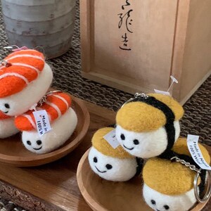 May include: Felt sushi keyrings in various styles, including nigiri and maki, with smiling faces. The keyrings are in orange, yellow, and white. They are presented on small wooden plates, with a wooden box in the background.