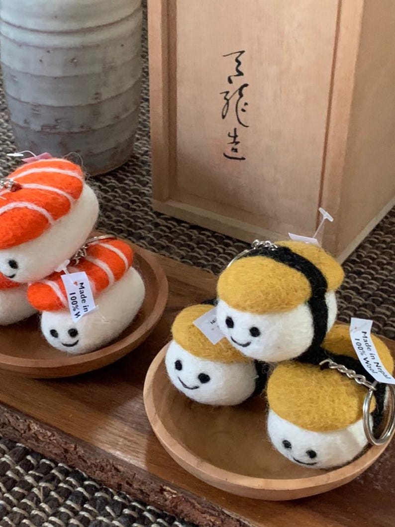May include: Felt sushi keyrings in various styles, including nigiri and maki, with smiling faces. The keyrings are in orange, yellow, and white. They are presented on small wooden plates, with a wooden box in the background.