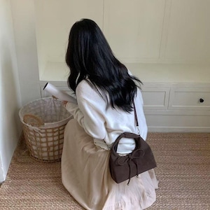 May include: A brown suede handbag with a rounded shape and a bow detail. The bag has a short handle and a long shoulder strap. The person is wearing a white sweater and a beige skirt. The background includes a woven basket and a neutral-toned rug.