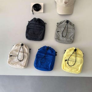 May include: A collection of drawstring backpacks in various colors, including black, gray, cream, blue, and yellow. A beige baseball cap with the text "ONEG Rew" is also displayed. The backpacks have a front pocket and black drawstring closure.