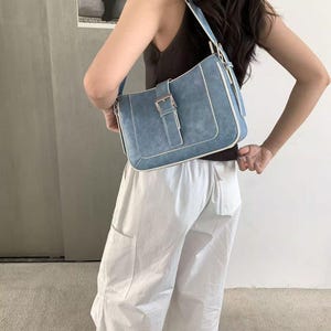 May include: A light blue shoulder bag with a rectangular shape and a buckle fastening. The bag has white trim and a matching strap. The bag is made of a suede-like material.