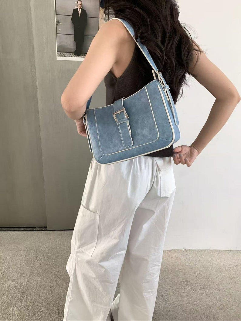 May include: A light blue shoulder bag with a rectangular shape and a buckle fastening. The bag has white trim and a matching strap. The bag is made of a suede-like material.