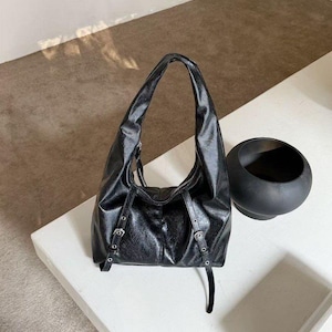 Puede incluir: Bolso de hombro de cuero negro con una sola asa. El bolso presenta hebillas plateadas y un dise&ntilde;o holgado. Un objeto decorativo negro y redondo se encuentra cerca sobre una superficie blanca.