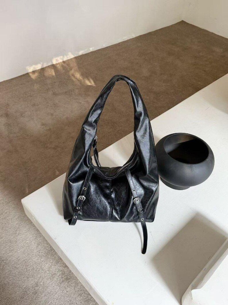 Puede incluir: Bolso de hombro de cuero negro con una sola asa. El bolso presenta hebillas plateadas y un dise&ntilde;o holgado. Un objeto decorativo negro y redondo se encuentra cerca sobre una superficie blanca.