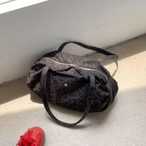 Puede incluir: Una bolsa de lona marr&oacute;n oscuro con estampado de leopardo, cremallera plateada y dos asas. La bolsa est&aacute; sobre una superficie de hormig&oacute;n junto a un par de zapatillas rojas con rayas blancas.