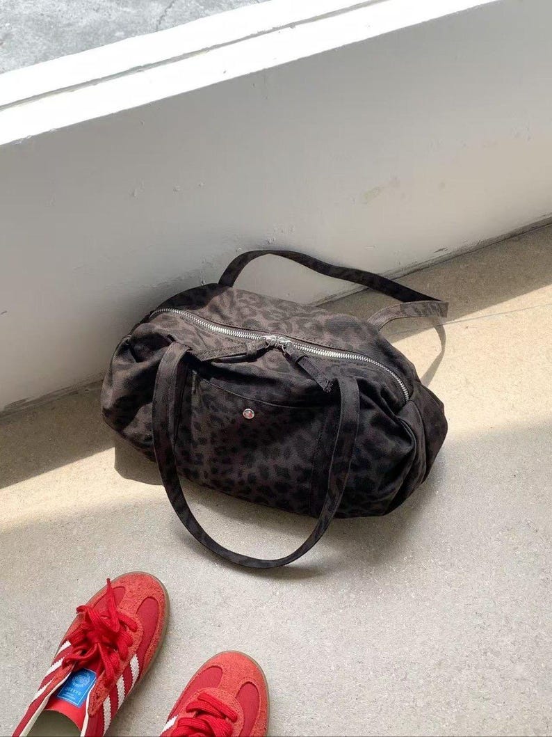 Puede incluir: Una bolsa de lona marr&oacute;n oscuro con estampado de leopardo, cremallera plateada y dos asas. La bolsa est&aacute; sobre una superficie de hormig&oacute;n junto a un par de zapatillas rojas con rayas blancas.