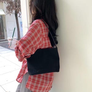 May include: A black rectangular shoulder bag with a thin strap. The bag is carried over the shoulder. The person is wearing a red and white plaid shirt and gray pants.