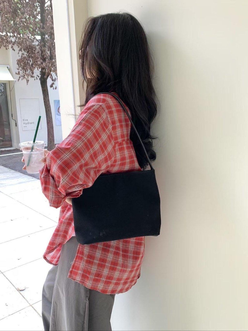 May include: A black rectangular shoulder bag with a thin strap. The bag is carried over the shoulder. The person is wearing a red and white plaid shirt and gray pants.